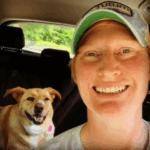 a woman wearing a baseball hat smiles at the camera with a dog smiling behind her in a car